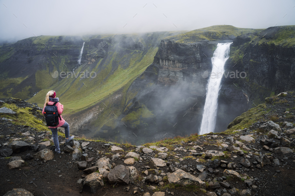 Woman with backpack and lilac jacket enjoying Haifoss waterfall of Iceland Highlands in - Stock Photo - Images