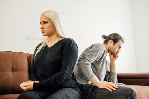 Stubborn angry husband and wife, spouses sit separate on sofa at home
