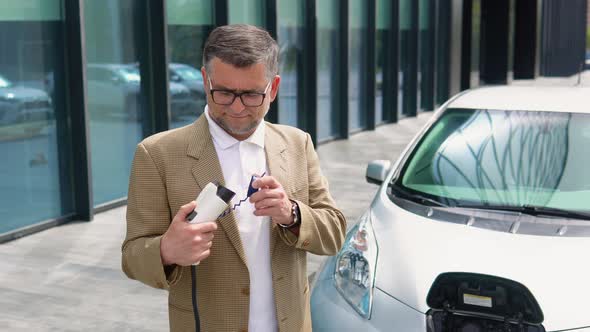 Older Man Stands with Charger Near His Electric Car alt