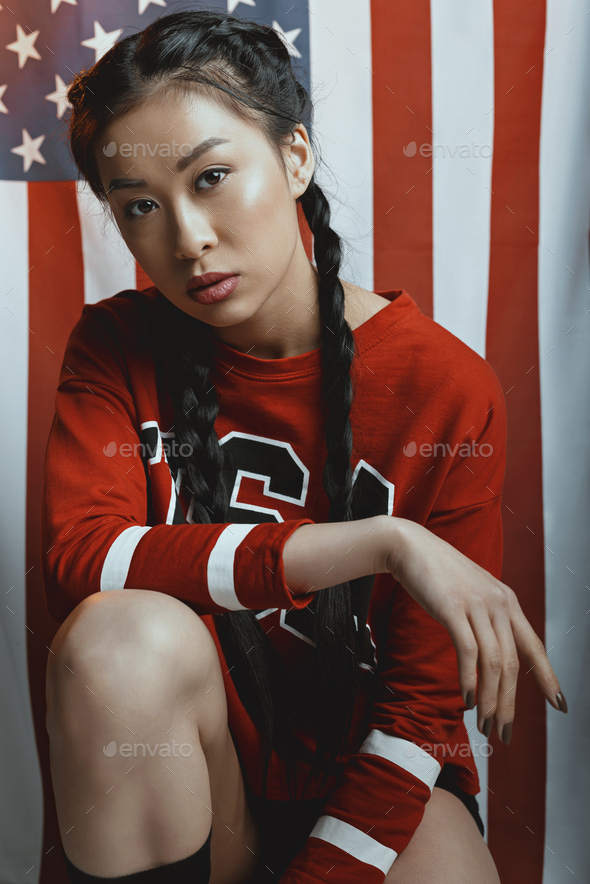 Hipster asian girl in american patriotic outfit with braids looking at camera with us flag on