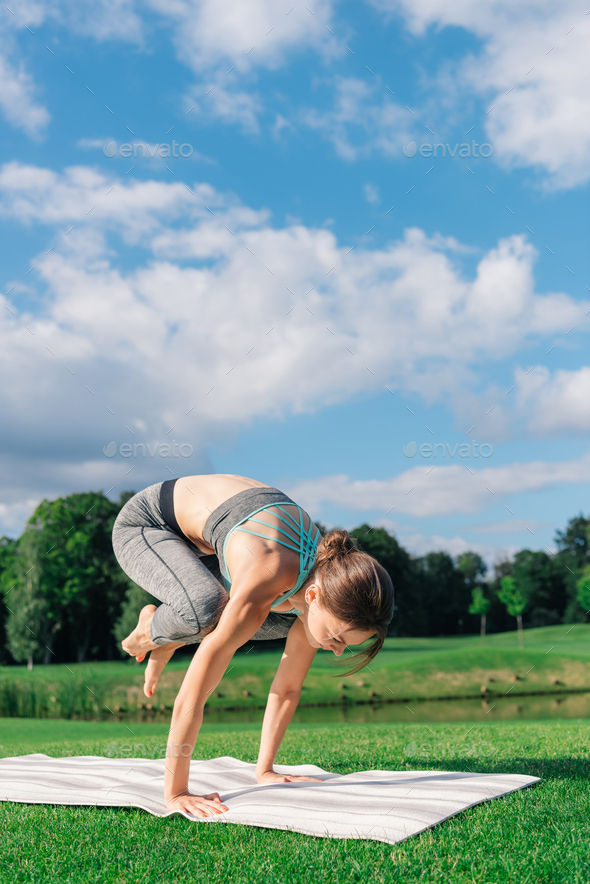 athletic young woman practicing yoga crow pose on yoga mat in park