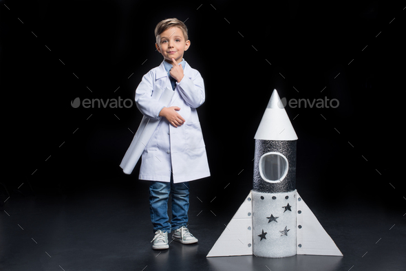 Little boy in white coat with finger on chin holding blueprints near toy rocket