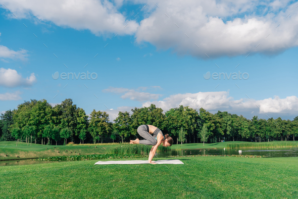 athletic young woman practicing yoga crow pose on yoga mat in park