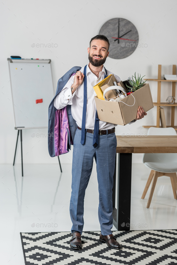 smiling businessman with cardboard box full of office supplies and jacket in hands quitting job