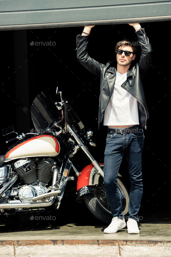 Handsome stylish young man in leather jacket and sunglasses standing in garage with motorcycle