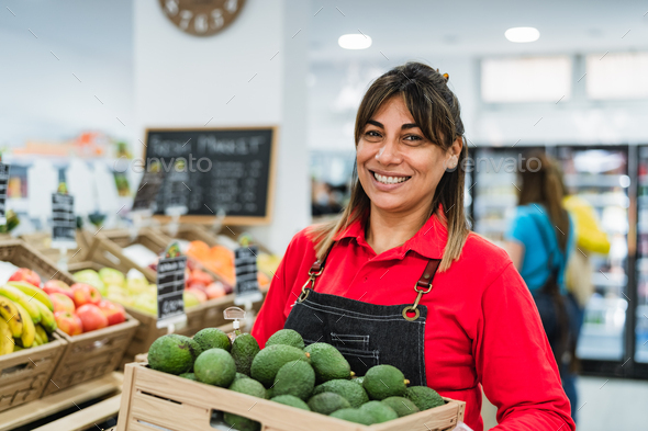 Latin woman working in supermarket holding a box containing fresh avocados