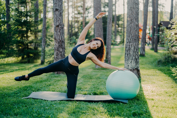 Active slim woman with cheerful expression leans aside does exercises with fitness ball