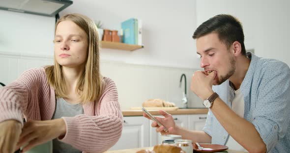 Young Couple Has Meal and Use Smartphones in Kitchen at Home. Woman Eats Chips