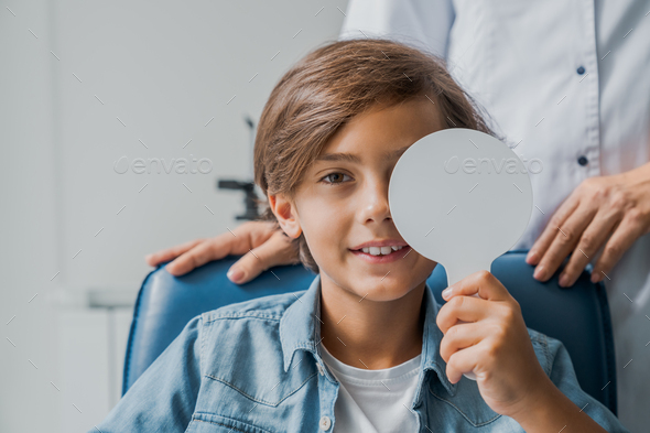 Smiling kid cover eye with medical tool for checking visual acuity in medical clininc cabinet