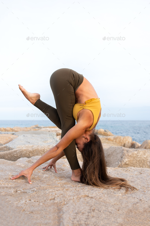 Woman doing yoga asana on a rock near the sea. Variation of forward fold / uttanasana. Vertical.