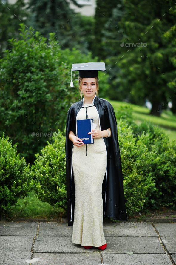 Portrait of a beautiful female graduate in dress and graduation gown with hat posing in the park.