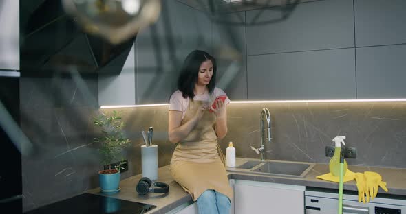 Lady Housewife Dressed in Apron Sitting on Kitchen Surface Use Smartphone After Cleaning at Home alt