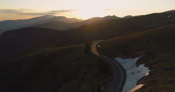Drone Follows Lonely Car Driving on Majestic Winding Mountain Road at Golden Sunset alt
