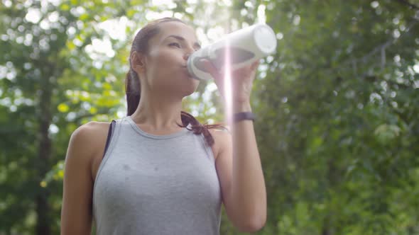Beautiful Sportswoman Drinking Water and Smiling at Camera in Park alt
