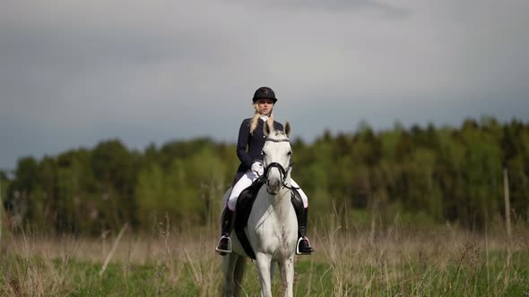 a Blonde in a Jockey Suit and a Riding Helmet Rides a White Horse Through a Field Against the alt