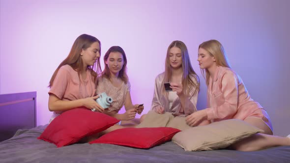 Four Happy Pretty Young Women in Pajamas Sits on Bed and Takes Selfies on Vintage Camera at alt