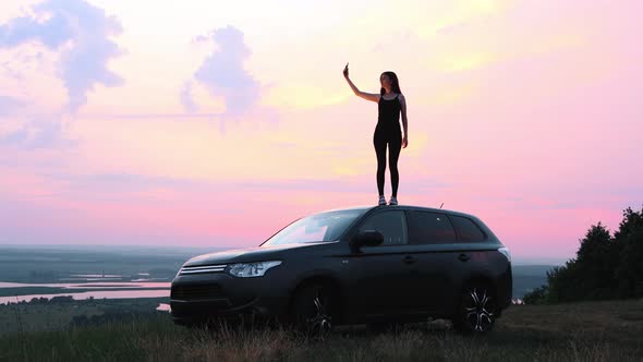A Young Woman Standing on the Roof of a Car and Trying to Find a Phone Connection alt