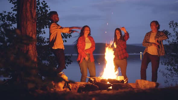 Excited Young Men And Women Hipsters Are Dancing Around Bright Campfire