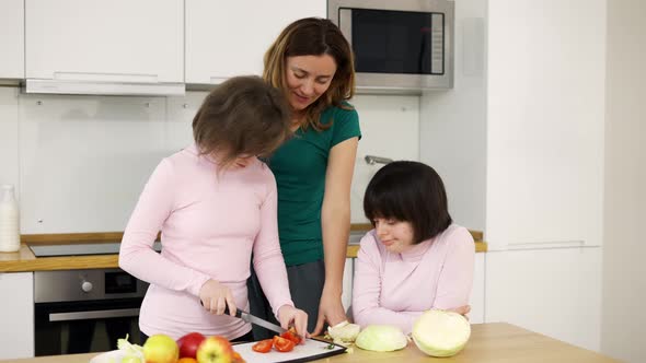 Two Girls with Down Syndrome Cutting Vegetables with Their Mother in Kitchen alt