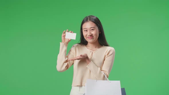 Asian Shopping Woman With A White Card And Shopping Bags Smile To Camera On Green Screen alt
