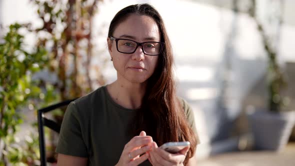 Portrait of Pretty Brunette Woman in Cafe Holding Smartphone Correcting Her Glasses alt