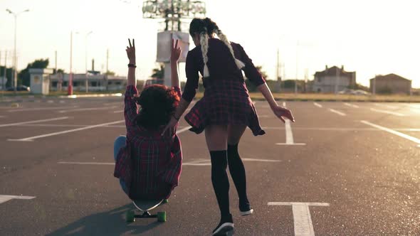 Cheerful Woman Sitting on a Longboard While Her Friend is Pushing Her Behind During Sunset alt