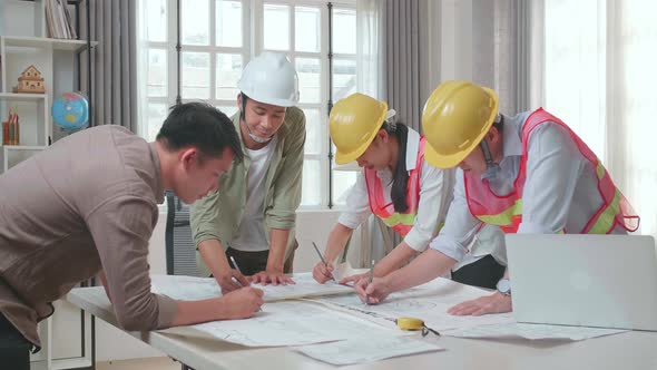 Three Asian Engineers With Helmets Helping A Man Drawing Building Construction At The Office alt