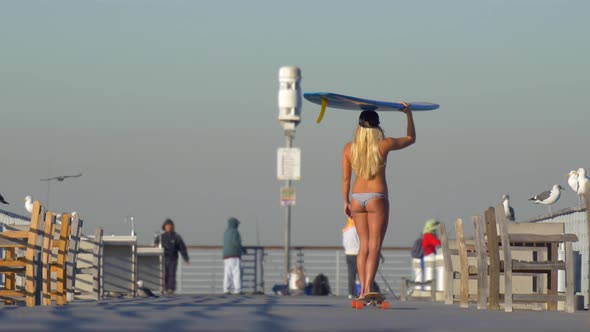 A young woman longboard skateboarding in a bikini while balancing a surfboard on her head. alt