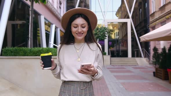 Woman in Modish Hat Walking Near Office Building with Street Cafe and Using Her Phone alt