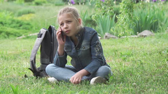 A girl of school age sits in a park on the grass and talks on the phone.