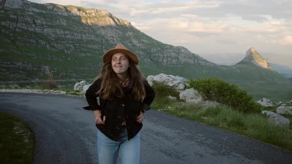 Smiling  hipster woman in hat with backpack along mountain landscape at sunset alt