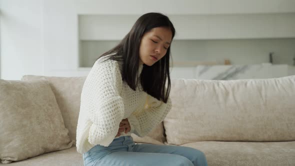 An Asian Woman in a White Blouse is Sitting on the Couch and Feels a Strong Abdominal Pain Which is alt