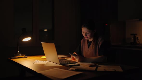 Beautiful Woman Working Late at Home Using a Laptop and Receiving Text Messages on Smartphone alt
