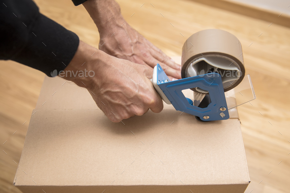 Hands of a man using a tape dispenser to seal a shipping box