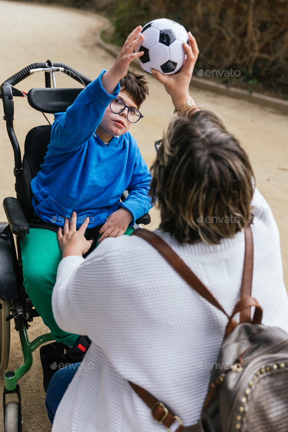 A mother playing with a soccer ball with her son with a disability in a wheelchair in the park.