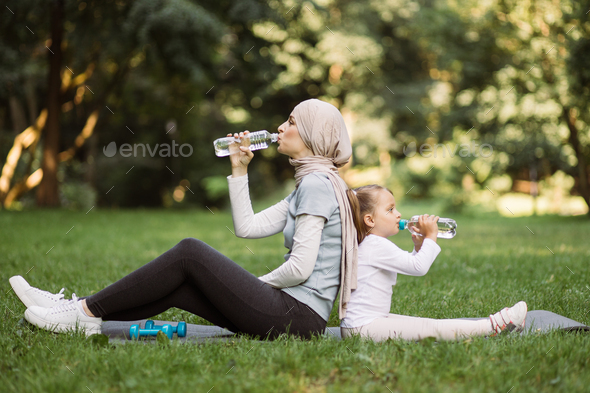 Muslim woman in hijab, sitting on mat with her little daughter at park, and drinking water