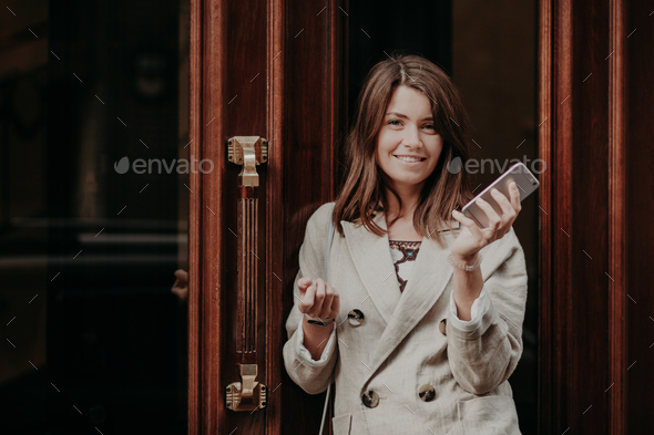 Elegant gorgeous lady in raincoat, holds mobile phone, waits for call, poses outdoor near doors.