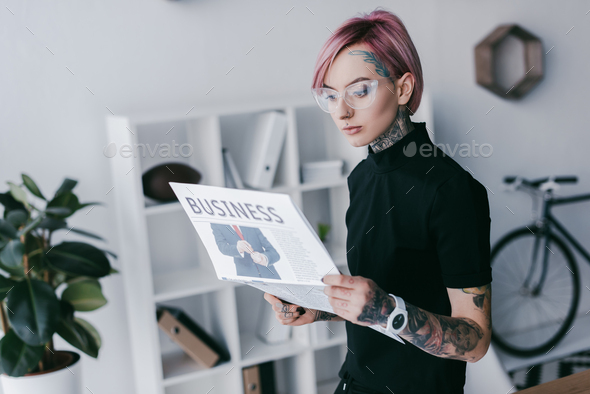 young tattooed businesswoman in eyeglasses reading newspaper at workplace