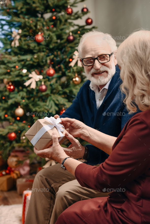 happy senior man presenting christmas gift to wife in front of beautiful christmas tree