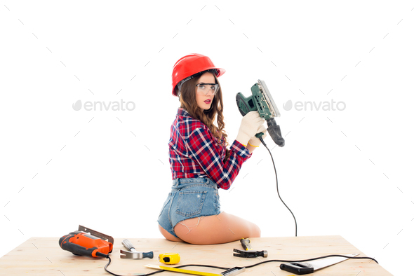 sexy girl in hardhat posing with grind tool on wooden table with tools, isolated on white