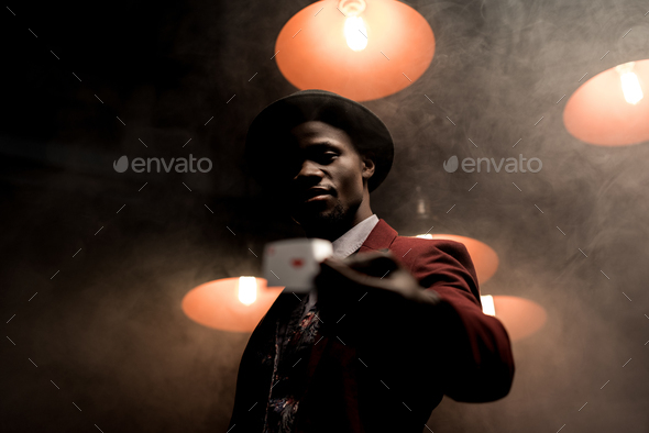 handsome lucky african american man with playing card in hands in dark smoky room with lamps