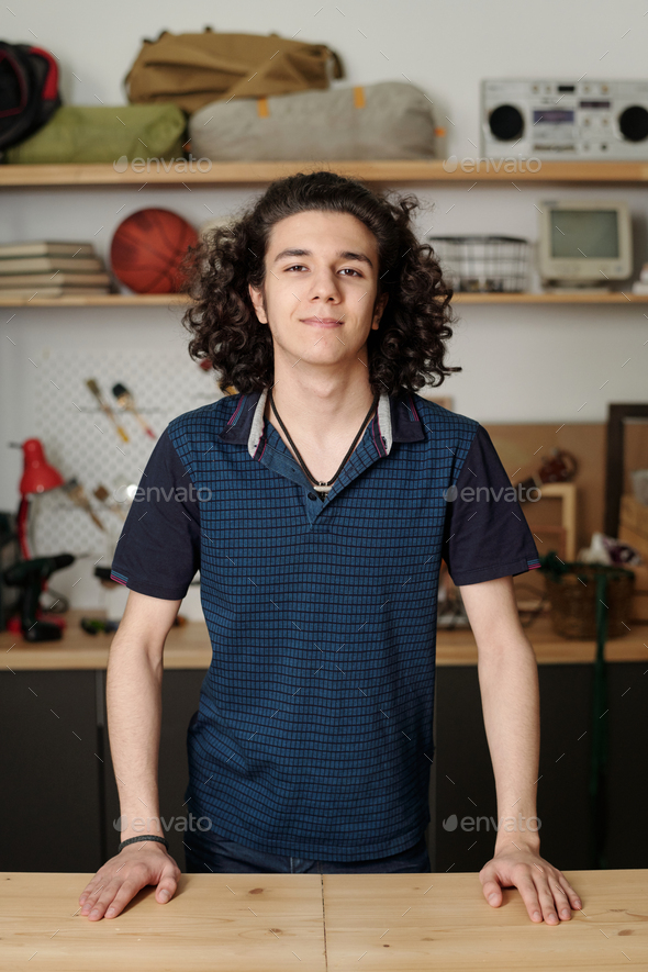 Happy adolescent guy in dark blue shirt standing by wooden table in garage