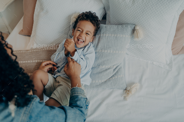 Sweet excited mixed race boy lying on bed and laughing while playing with his loving mom