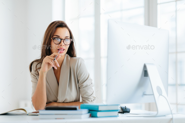 Contemplative female entrepreneur keeps pen in mouth, focused in monitor of computer.