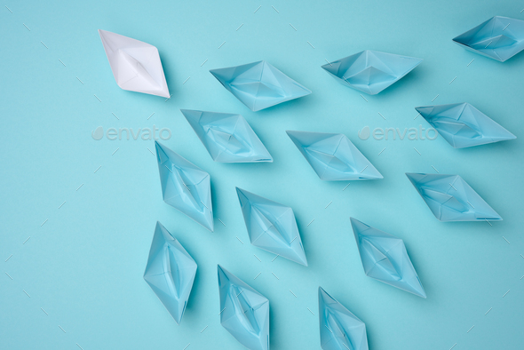 Group of blue paper boats follow white against a light blue background