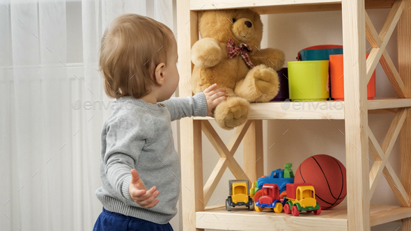 Cute baby boy taking teddy bear toy from bookshelf in playroom. Child education - Stock Photo - Images