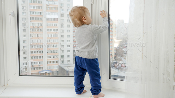 Little baby boy standing on windowsill and pulling window handle. Baby in danger. CHild safety. - Stock Photo - Images