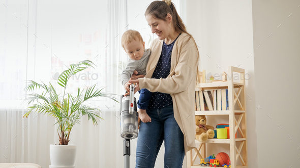 Young mother cleaning floor with vacuum cleaner while holding her little baby son. - Stock Photo - Images
