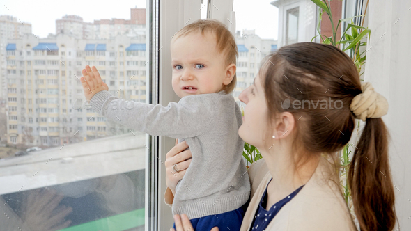 Young mother holding her baby son trying to open window on high floor. Baby in danger. Child safety - Stock Photo - Images
