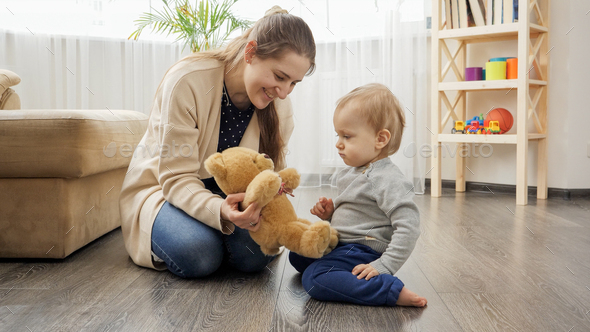 Happy smiling mother playing with her baby son with teddy bear on floor in living room. - Stock Photo - Images